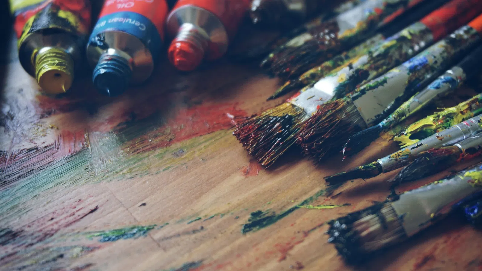 Assorted paint tubes and brushes on a wooden surface, showcasing vibrant colors and textures.