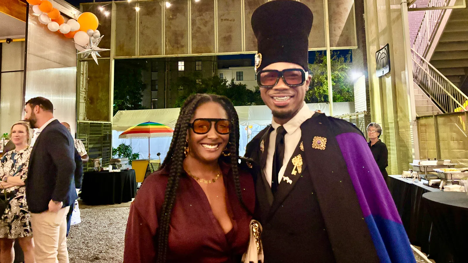 Two stylish people smiling at an outdoor nighttime event with decorative lights and party decorations.