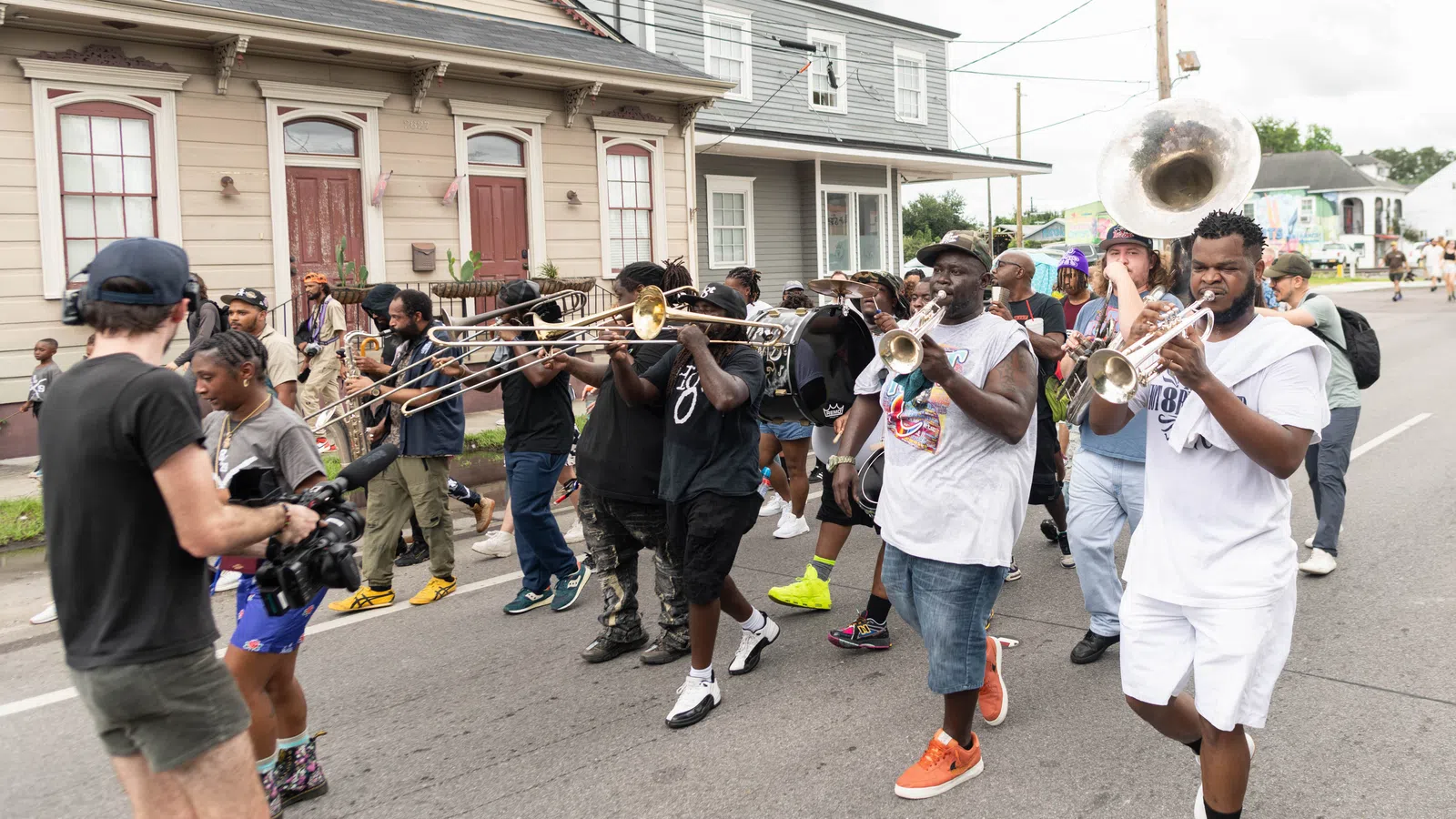 Street parade with musicians playing brass instruments and a cameraman filming the lively event.