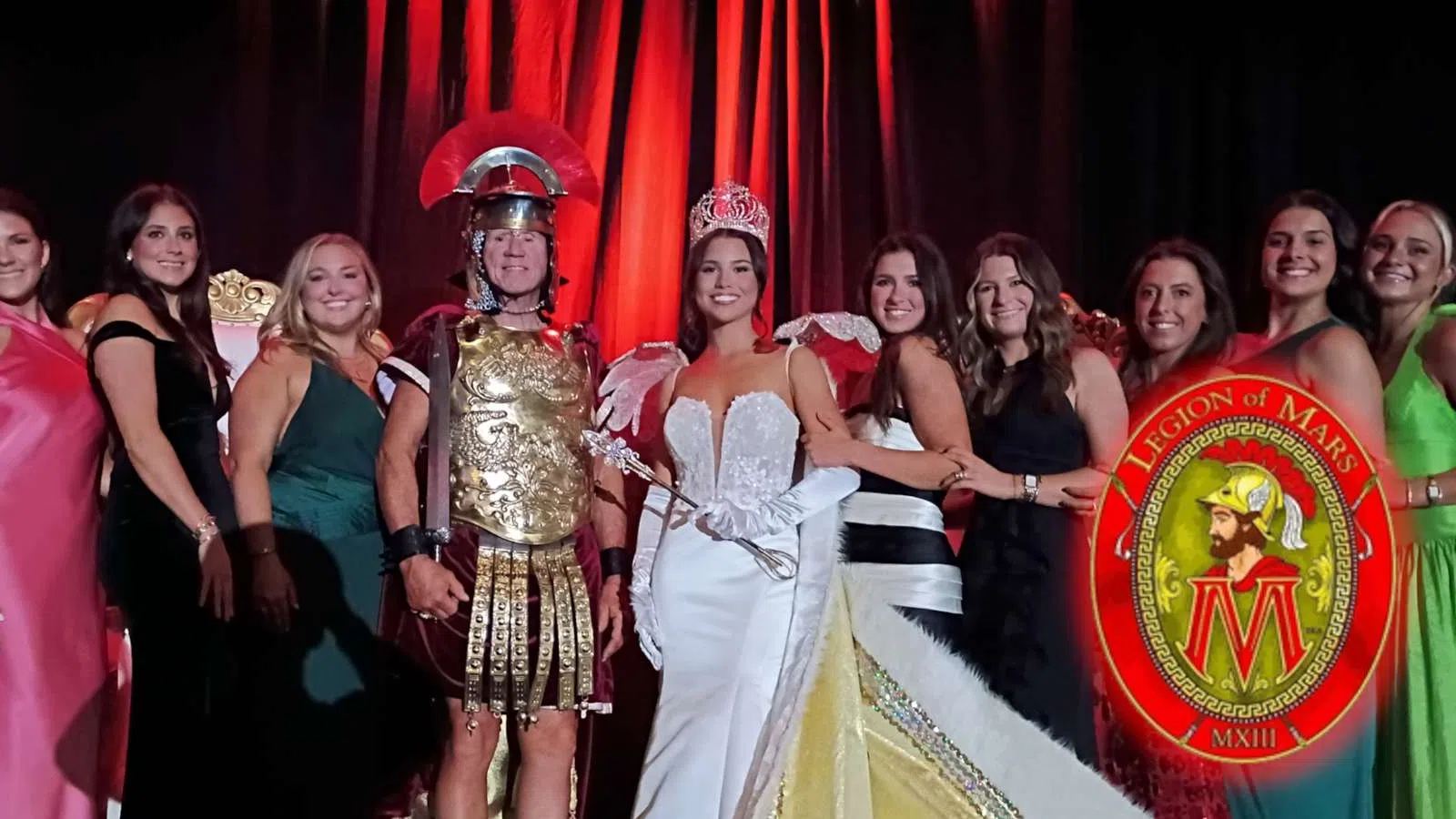 Group of people dressed formally with one man in Roman soldier costume and a crowned woman in white gown.