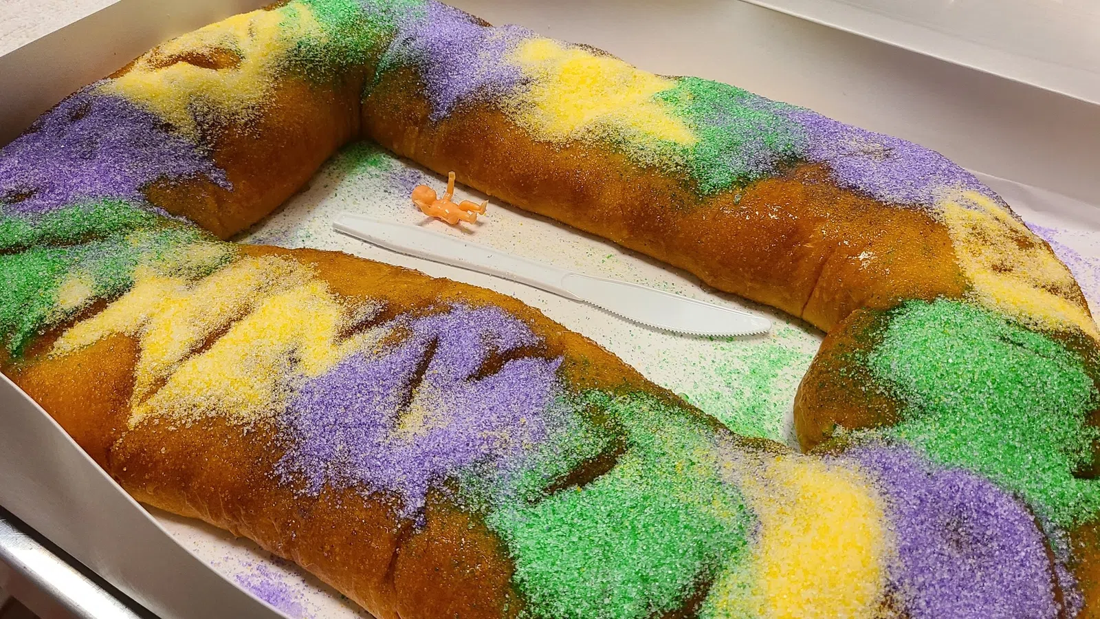 Traditional King Cake decorated with purple, green, and gold sugar in a white box with a plastic knife