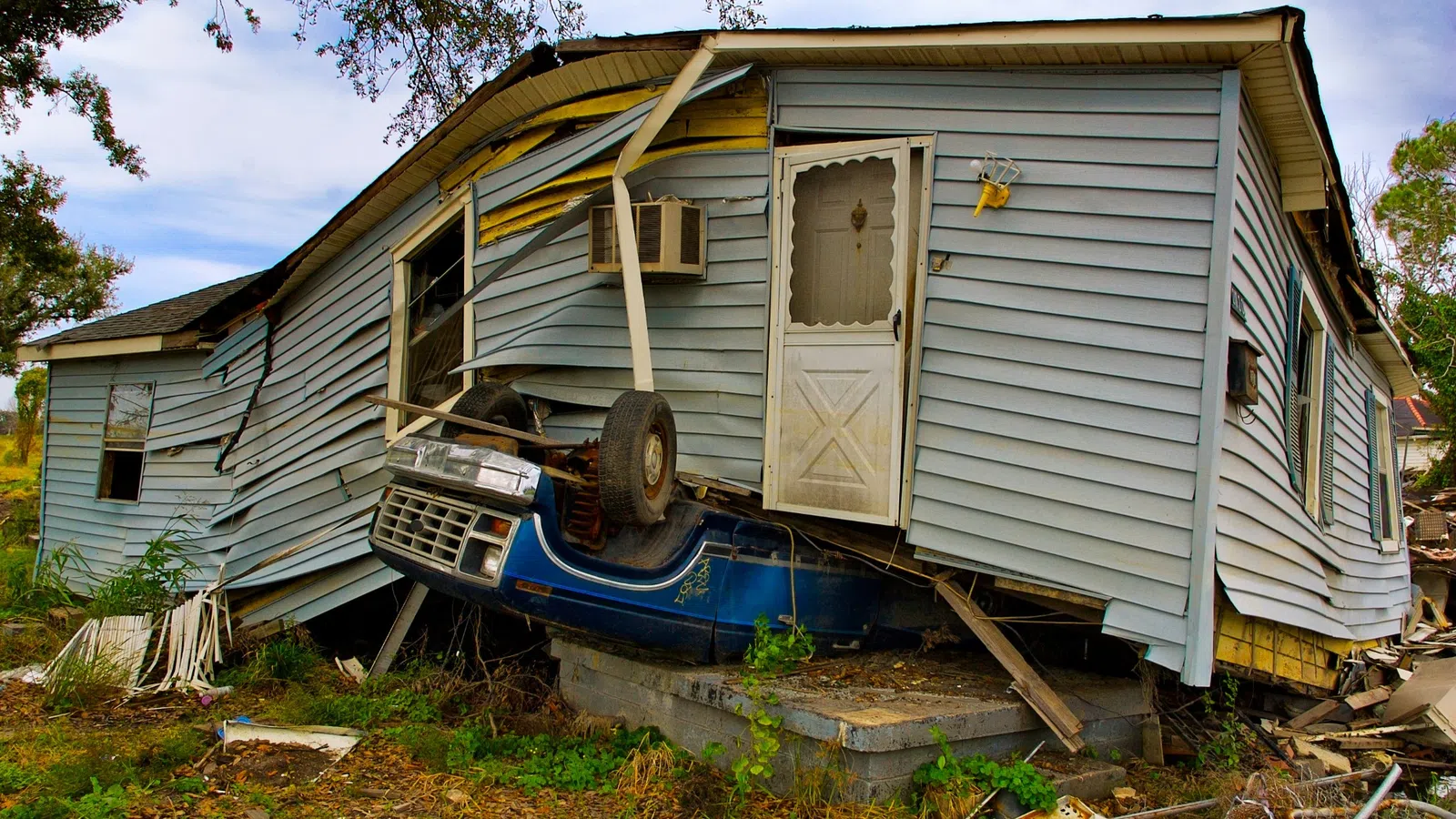 A damaged house with the front lifted by a blue car crushing the foundation amid debris and overgrown grass.