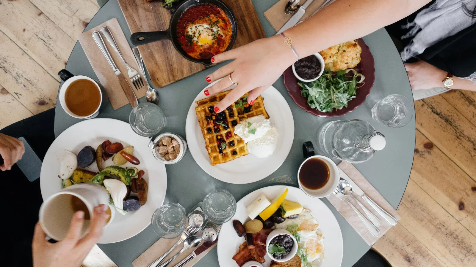 Overhead view of a breakfast table with waffles, eggs, coffee, salad, and people reaching for food.