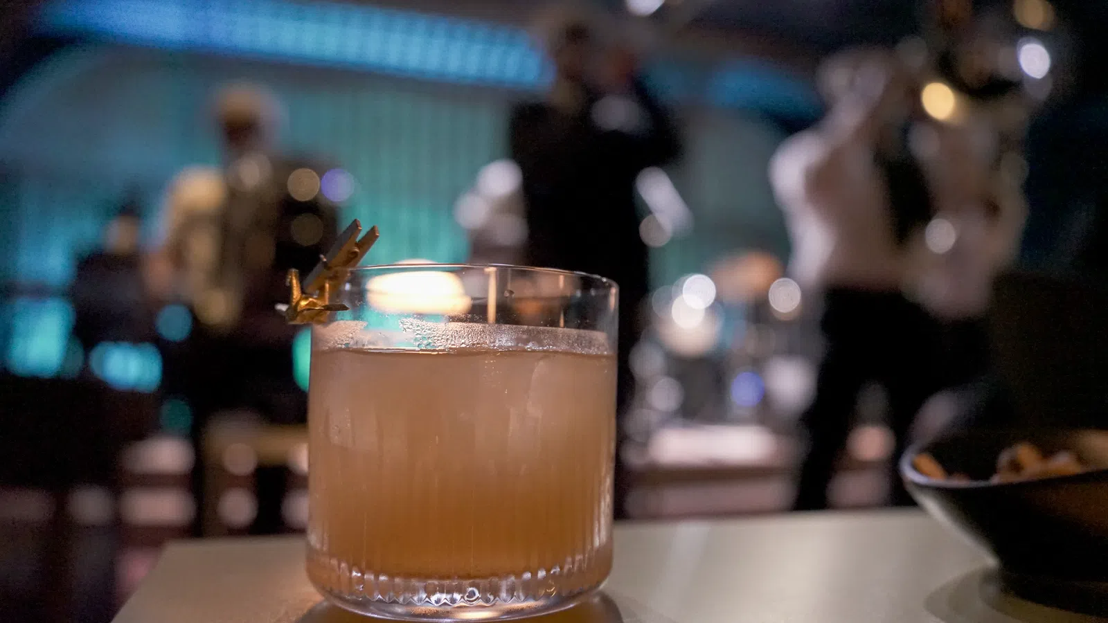Cocktail glass with an orange drink and garnish on a table in a dimly lit jazz bar with musicians playing in the background