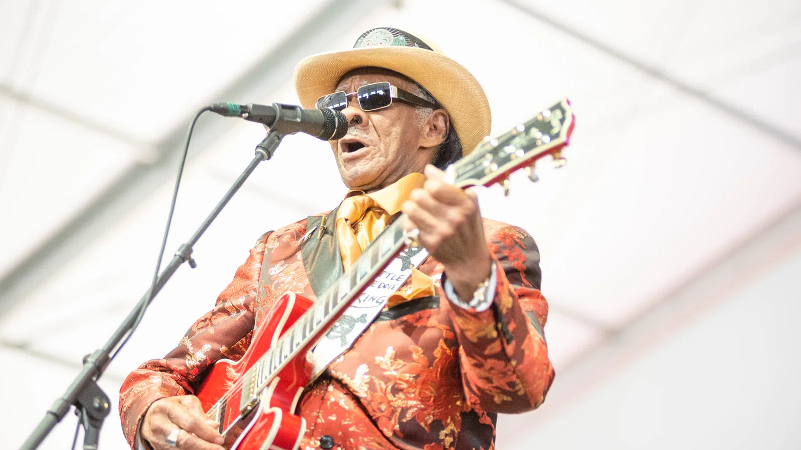 Musician in floral suit and hat playing electric guitar and singing into microphone on stage.