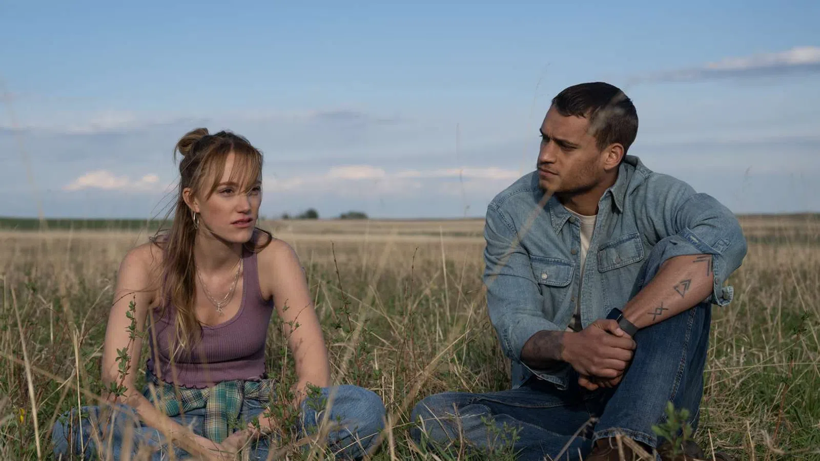 Young man and woman sitting in a grassy field talking under a partly cloudy sky on a sunny day