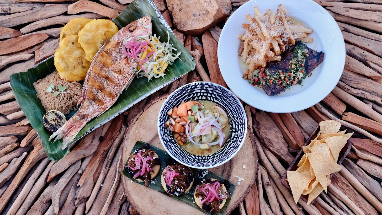 Assortment of grilled fish, fries, tostones, plantain chips, and small appetizers served on wooden and banana leaf platters.