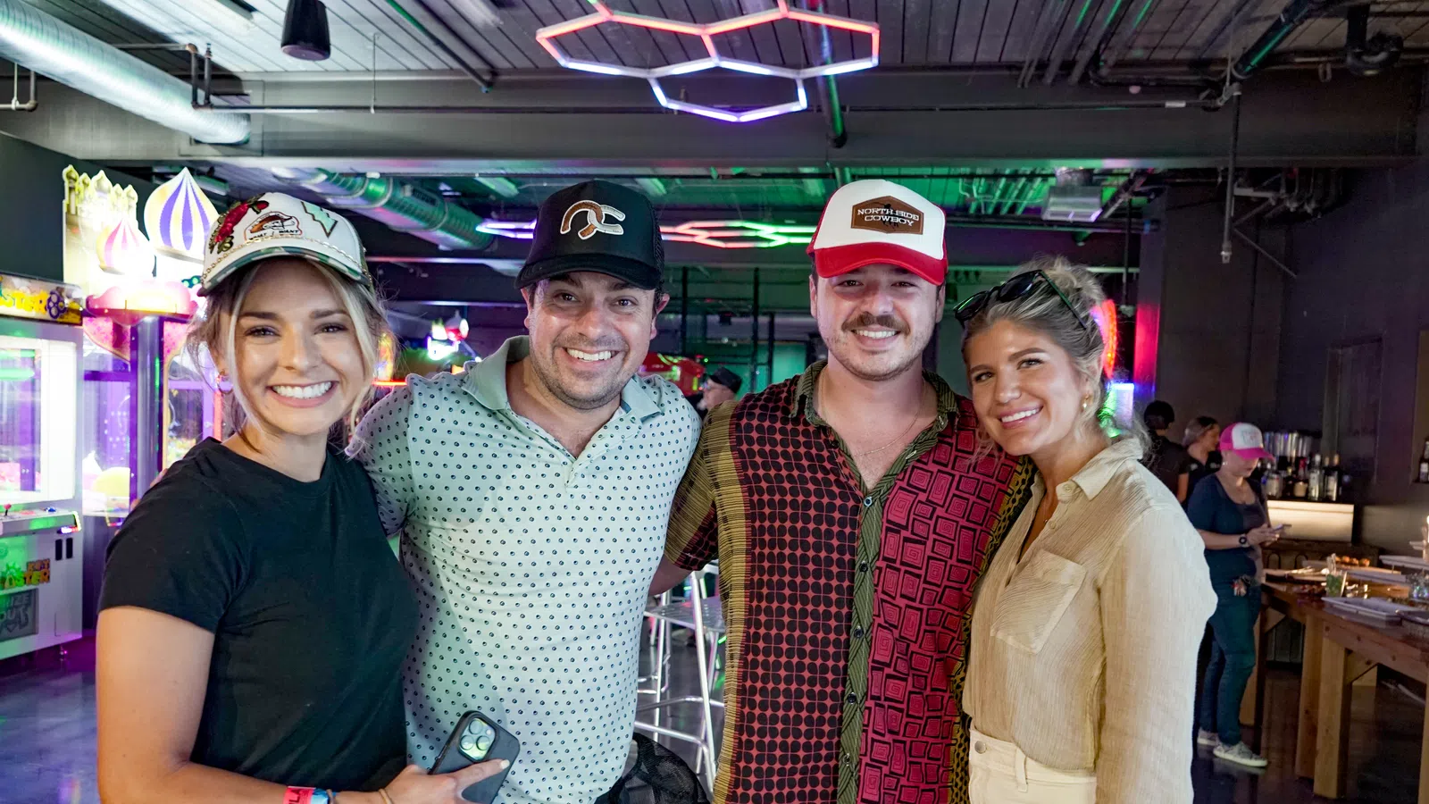 Four friends smiling in a vibrant arcade with colorful neon lights and casual attire including hats and wristbands.