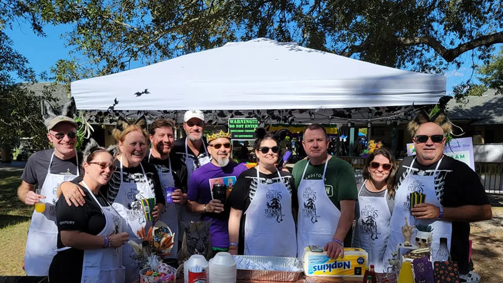 Group of ten people wearing aprons posing behind a decorated table at an outdoor event under a white canopy tent.