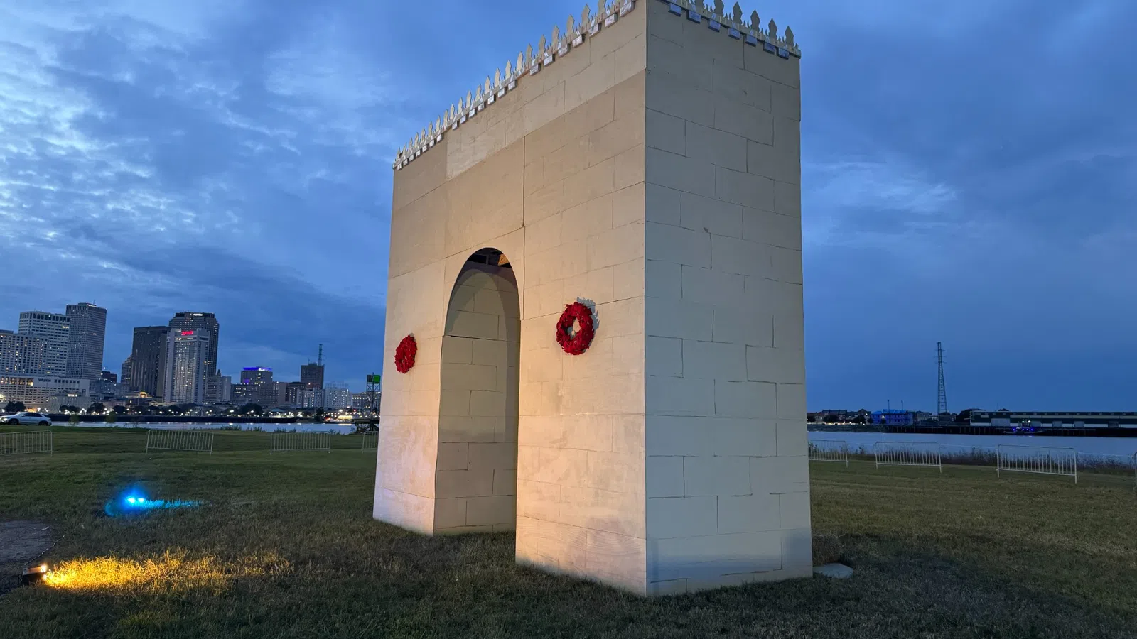 Large white stone monument with red wreaths on a grassy field at dusk with a city skyline in the background
