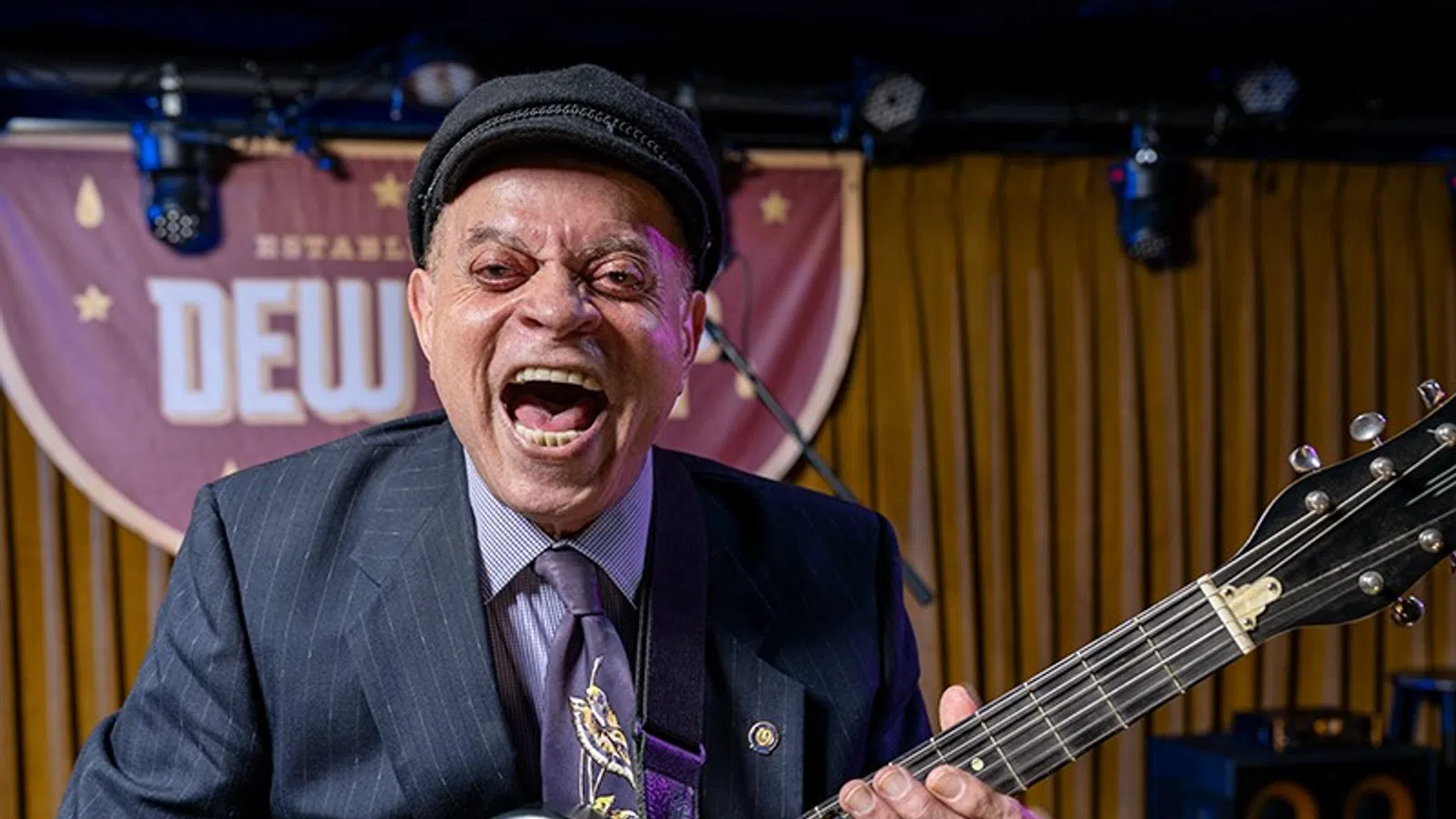 Smiling man in a suit and hat playing electric guitar on a stage with wooden backdrop and lights