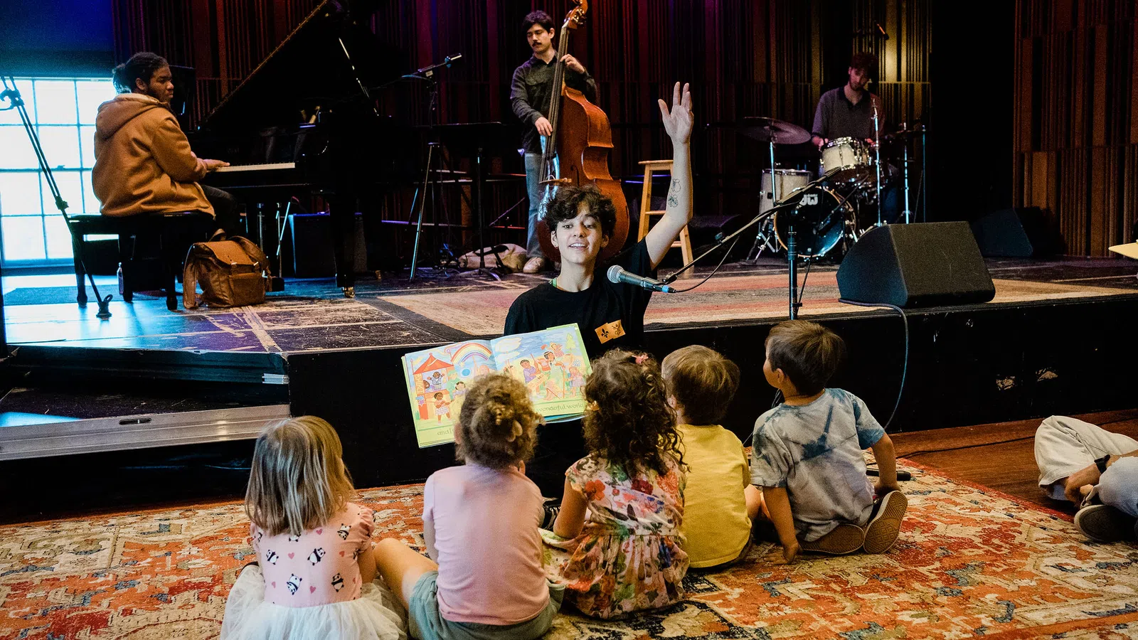 Adult reading a colorful book to children seated on a rug with live musicians performing in the background
