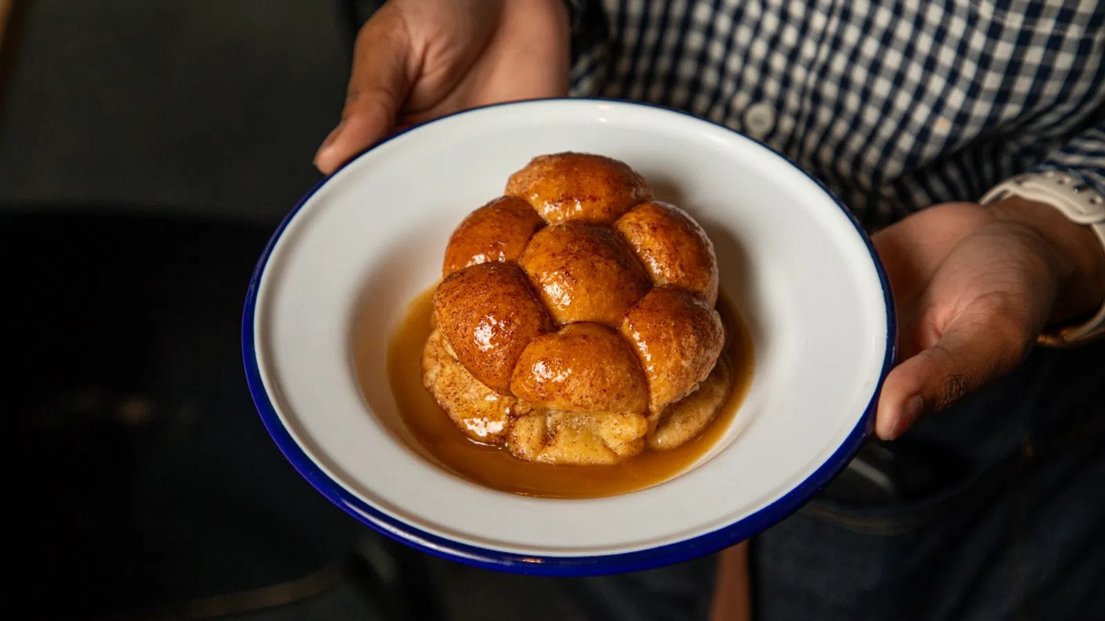 Person holding a white plate with golden brown sticky buns drizzled in syrup inside a rustic setting