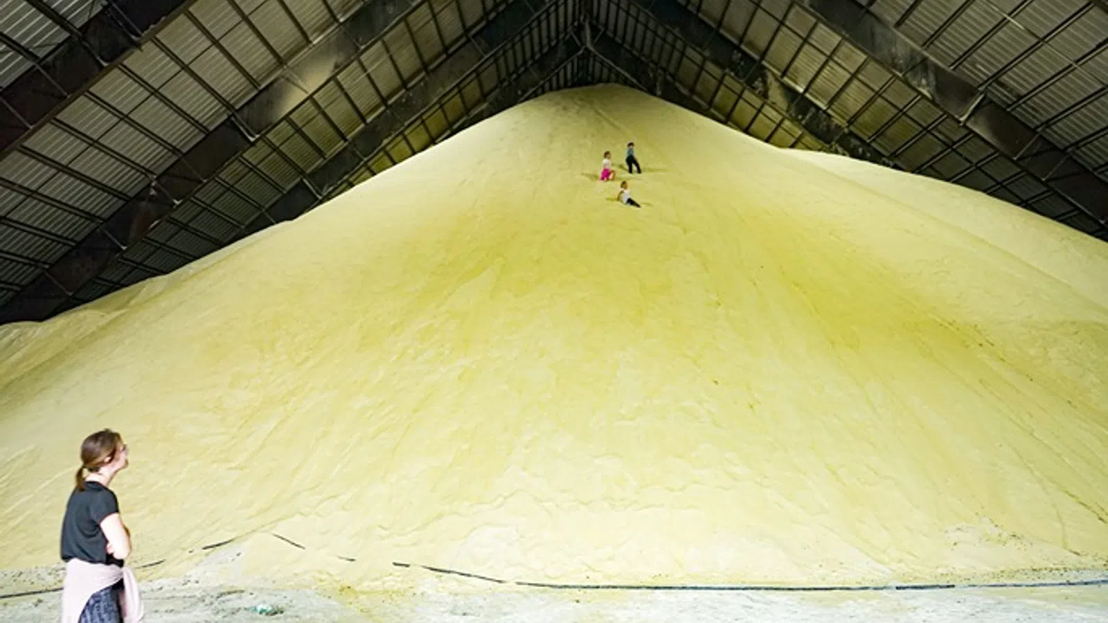 Children play on a large yellow sulfur pile inside an industrial warehouse with a woman watching.