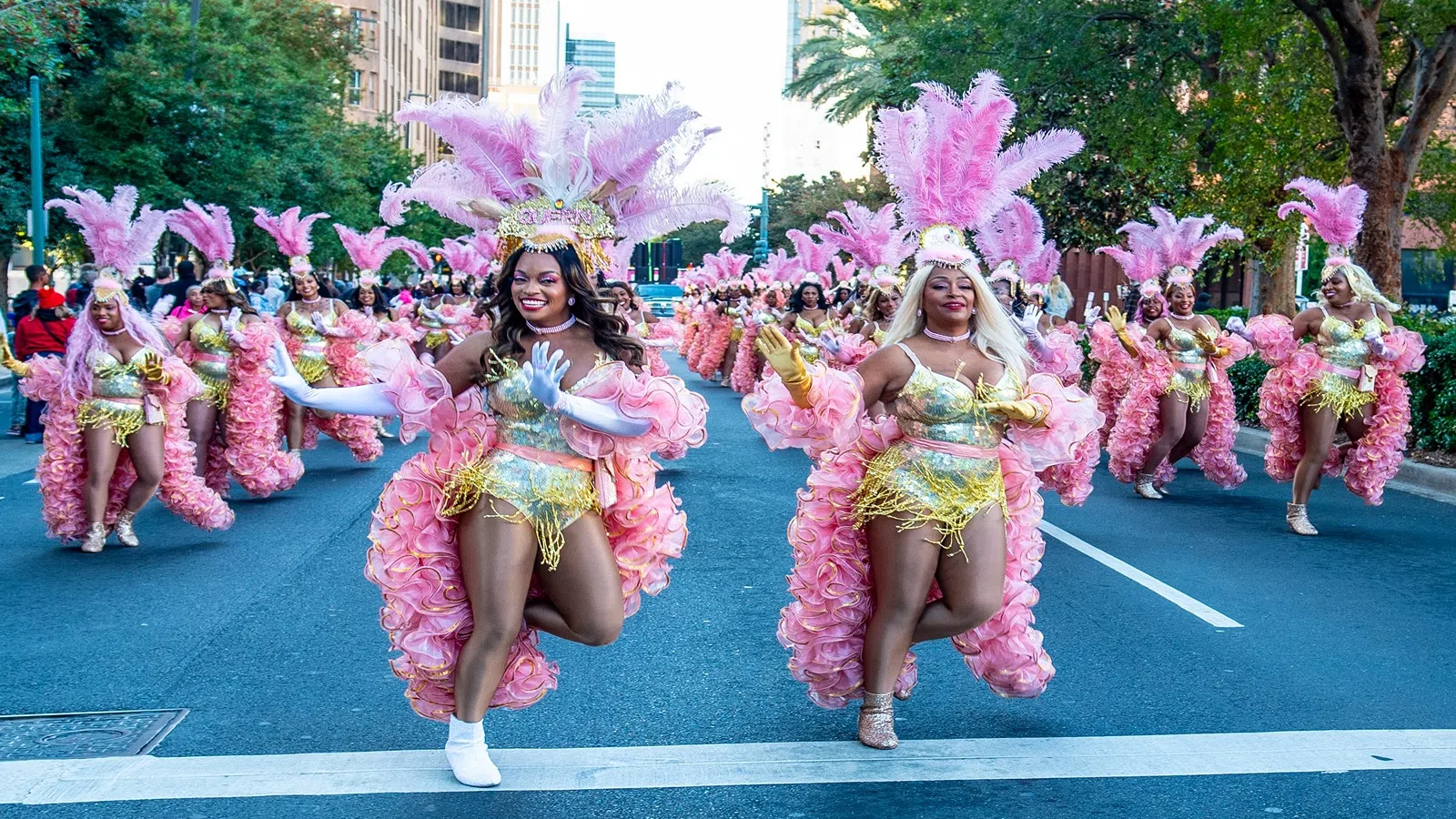 Dancers in pink feathered costumes perform in a vibrant street parade in a city setting, smiling and energetic.