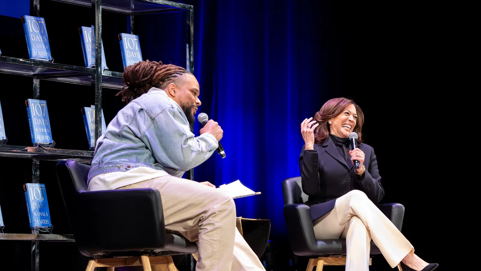 Two people on stage during a lively interview with books displayed on shelves and blue curtains in the background