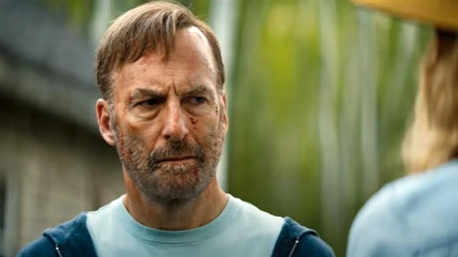 Intense bearded man with facial cuts looks serious outdoors with blurred greenery and person in foreground