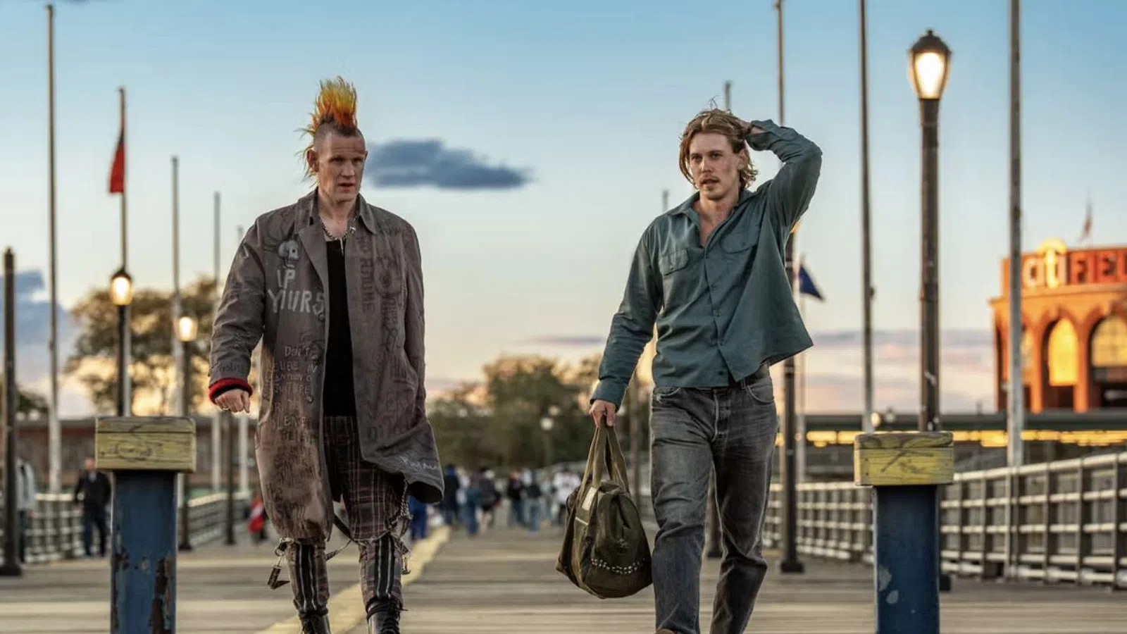 Two young men walking on a wooden pier at sunset, one with a colorful mohawk and punk style, the other casual with a bag.
