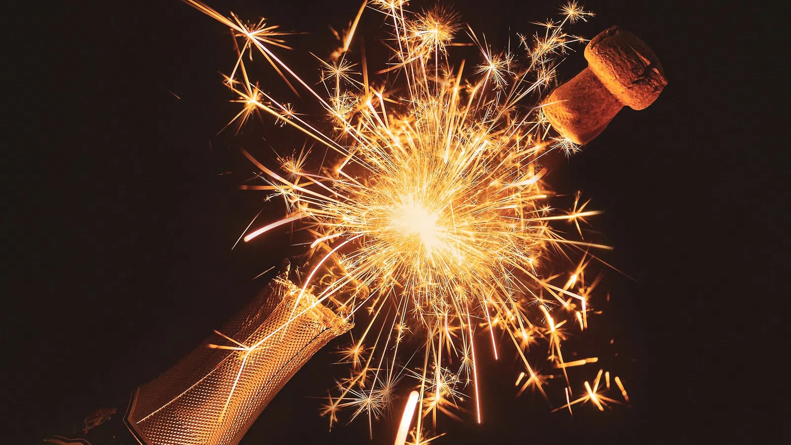 Champagne bottle popping with sparkling fireworks bursting from the neck against a dark background.