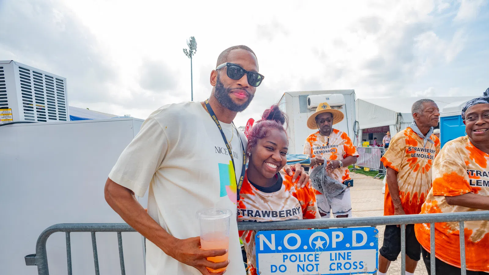 A smiling couple at a festival pose near a police barrier with festival staff in vibrant shirts in the background.