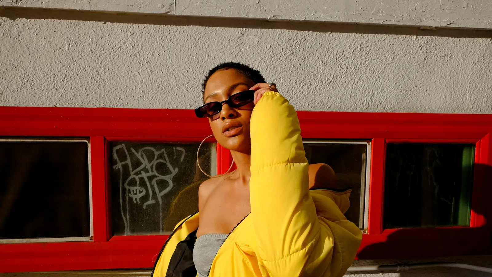 Woman in a bright yellow puffer jacket and sunglasses poses confidently against a textured wall with red window frames.