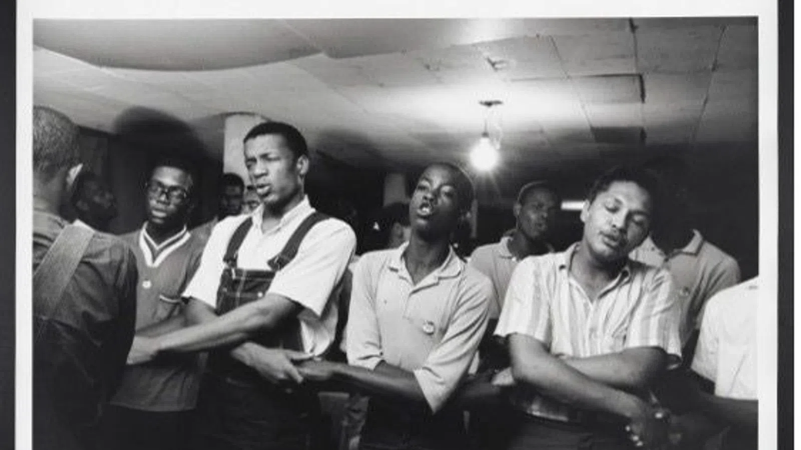 Black and white photo of young men standing with arms linked in a room with exposed ceiling light.
