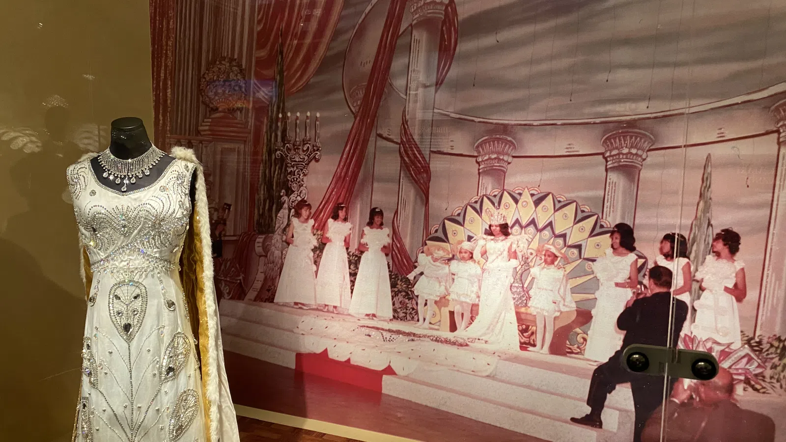 Vintage beaded white gown displayed next to a stage backdrop with women in white dresses during a pageant.