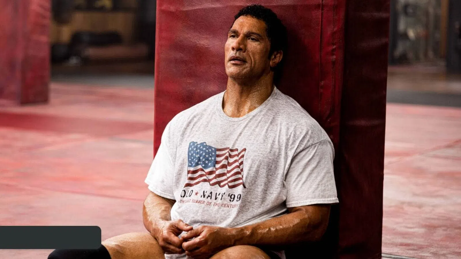Man in a white USA flag T-shirt resting against gym equipment in a workout space