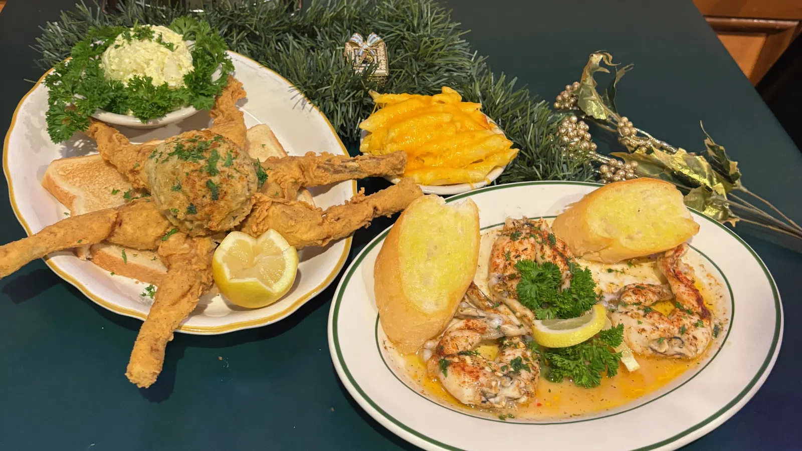 Plates of stuffed crab claws with toast, cocktail sauce, and macaroni and cheese on a green table with decorative greenery.