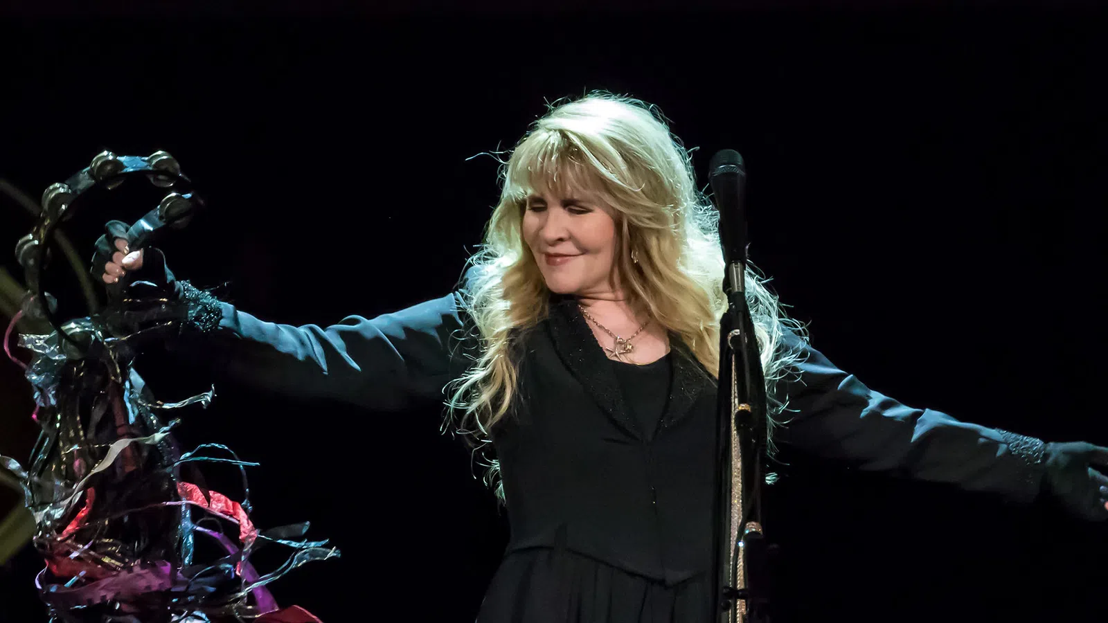 Female singer with long blonde hair performs on stage holding decorated microphone stand under spotlight.
