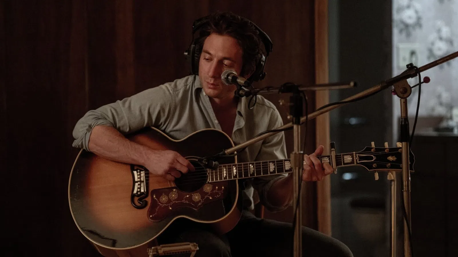 Man wearing headphones playing an acoustic guitar and singing into a microphone in a recording studio.