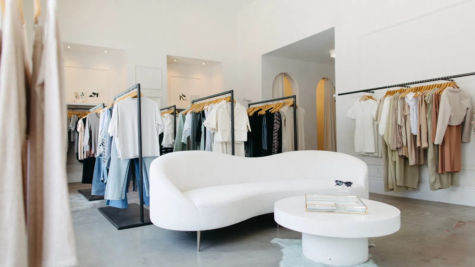 Modern boutique interior with white curved sofa, round table, and racks of stylish clothing on white walls.