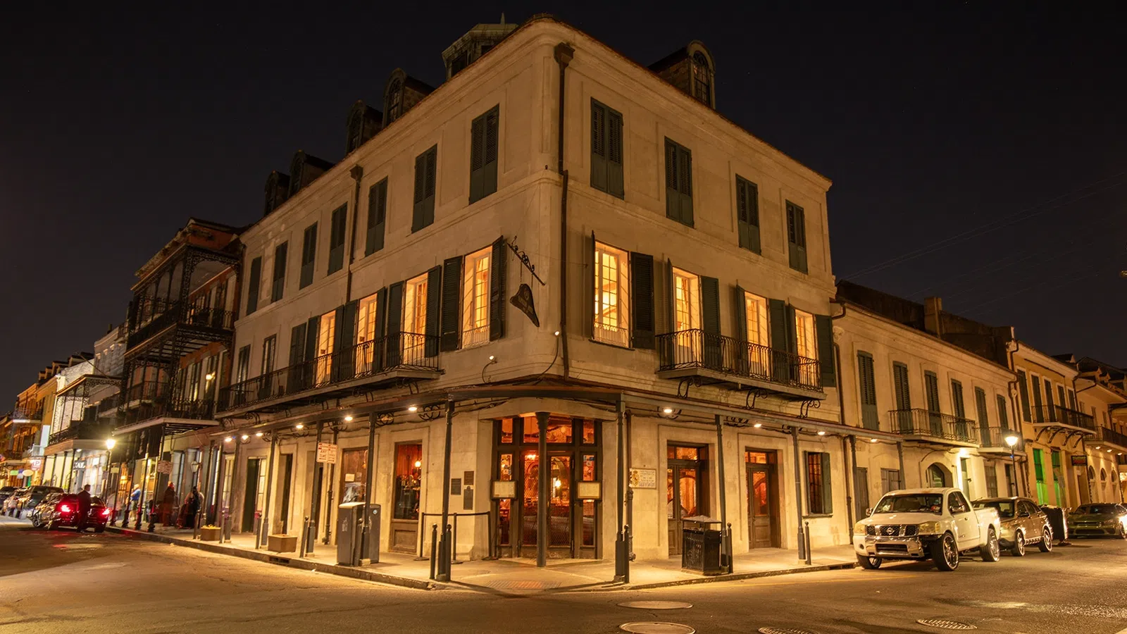 Historic corner building at night with warm lit windows and parked cars on street in a lively city area