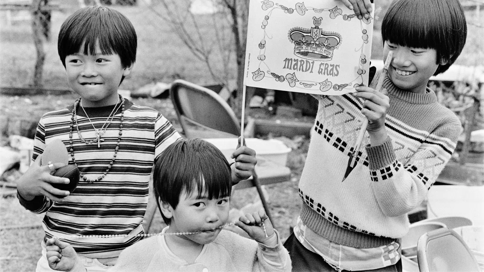 Three children smiling and playing outdoors, one holding a Mardi Gras flag, others wearing necklaces and festive clothes.