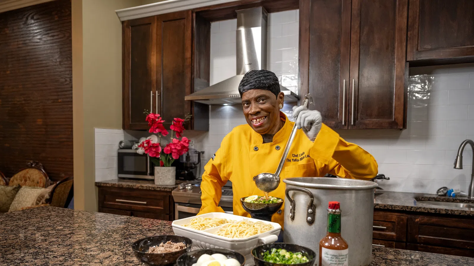Chef in an orange shirt prepares dishes in a modern kitchen with fresh ingredients and vibrant decor.