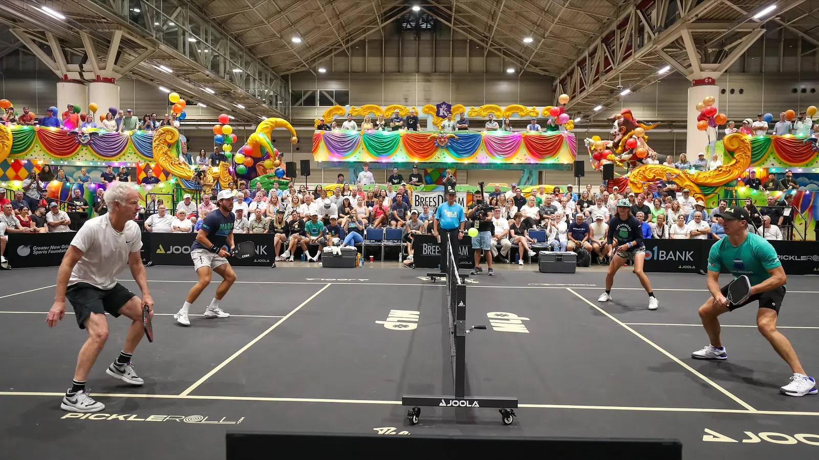 Indoor pickleball doubles match with players poised at the net and a large colorful audience in the background