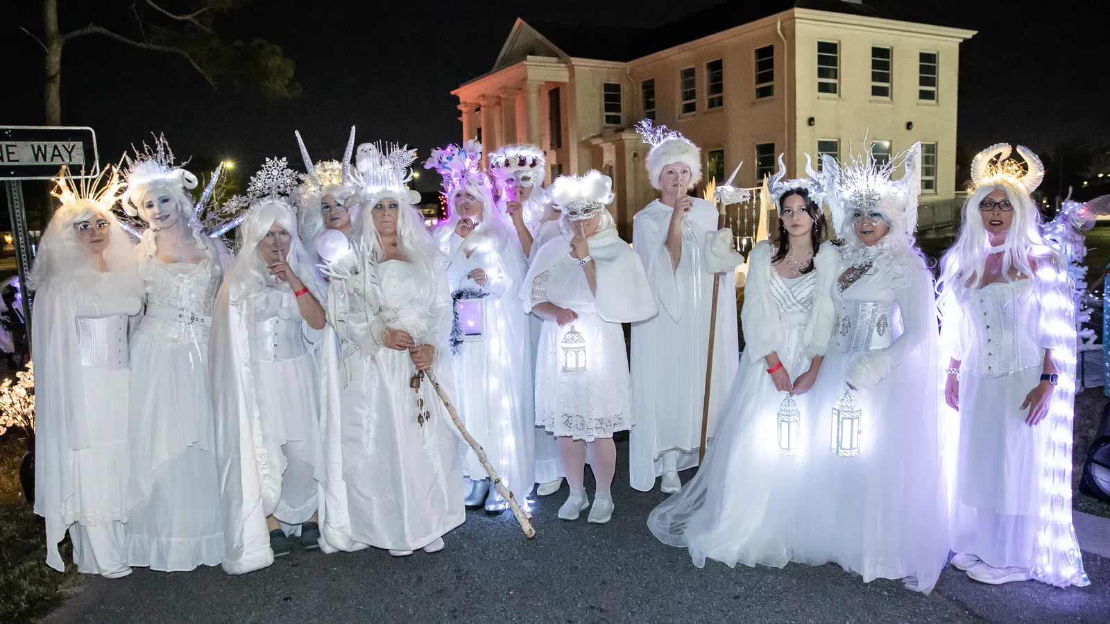 Group of people in elaborate white costumes and glowing accessories posing outdoors at night near a building