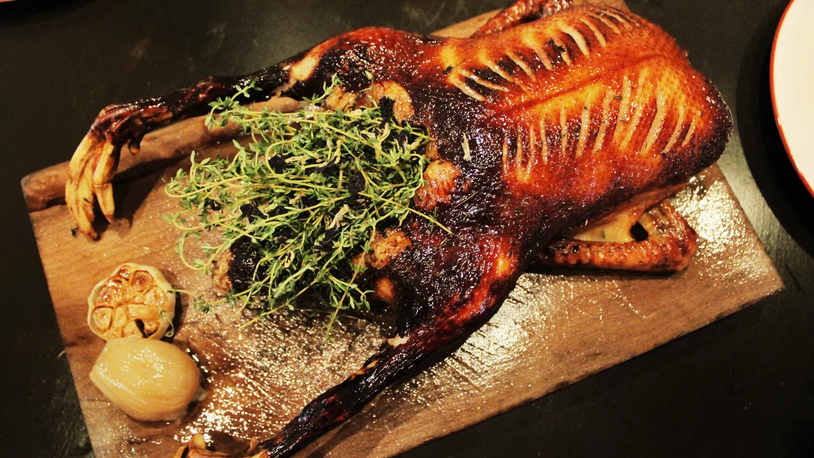 Whole roasted duck with crispy skin garnished with fresh herbs served on wooden board with garlic and onion.