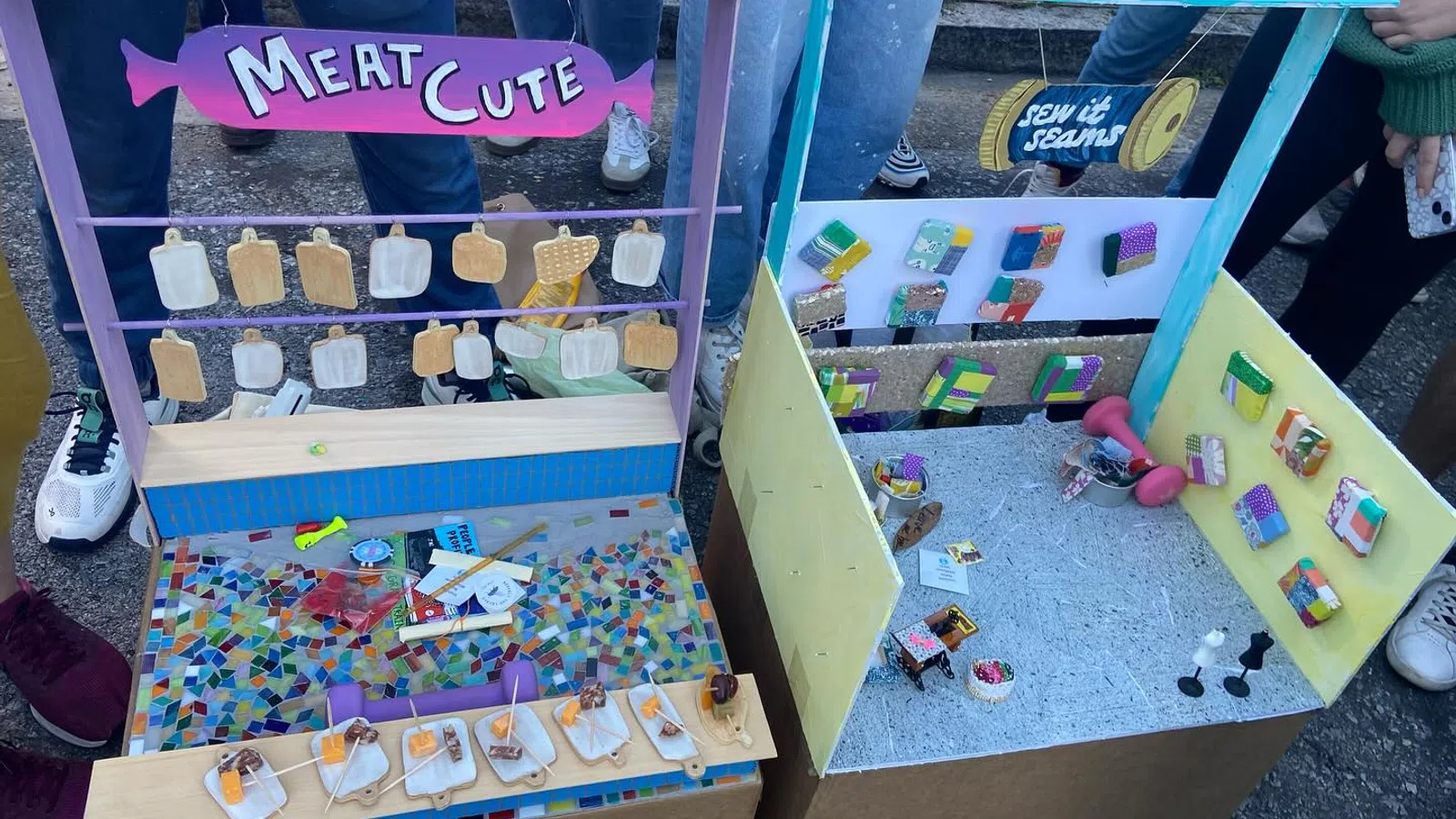 Two colorful miniature market stalls with handmade items labeled Meat Cute and Sew It Seams displayed outdoors with people nearby.