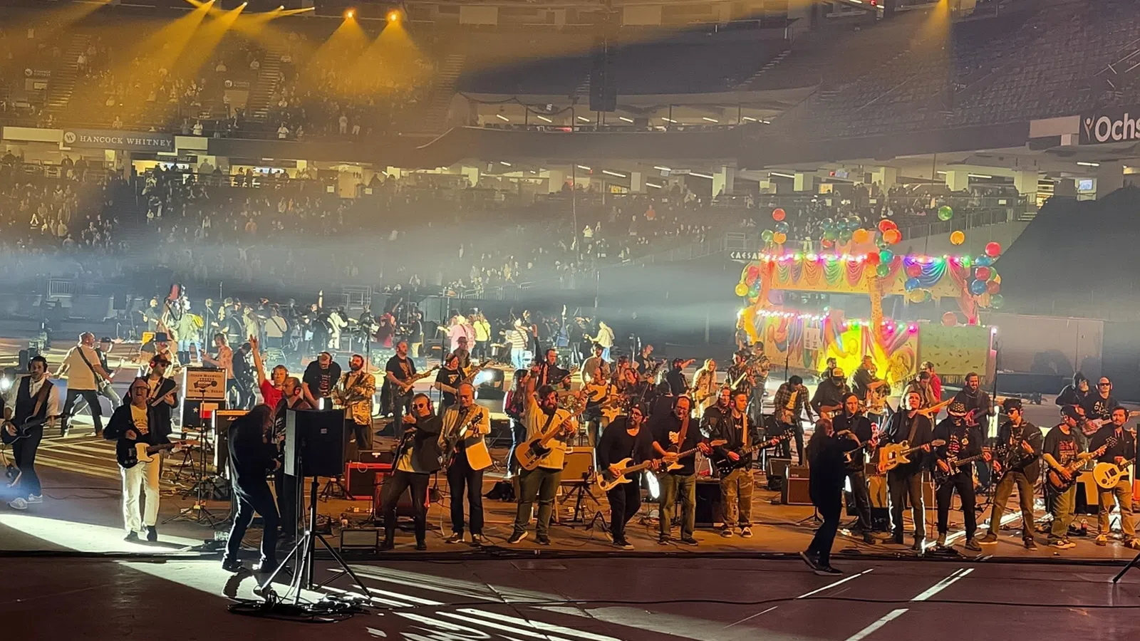 Large group of musicians performing on stage in a stadium with colorful lights and audience in background