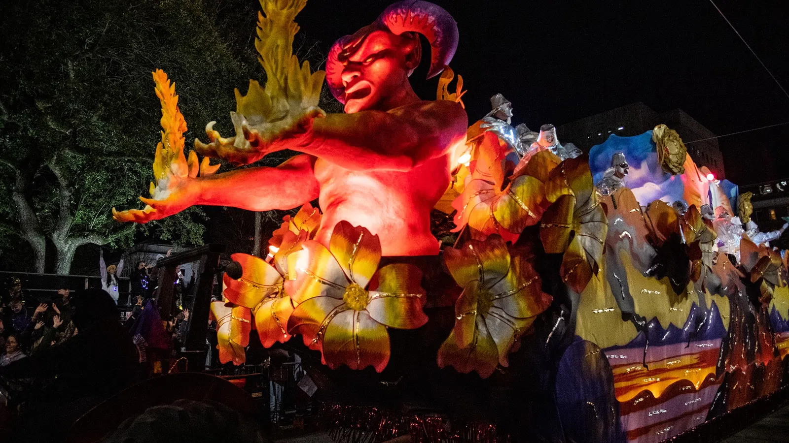 Illuminated red demon float with yellow flowers and flames at a night parade with spectators in background.