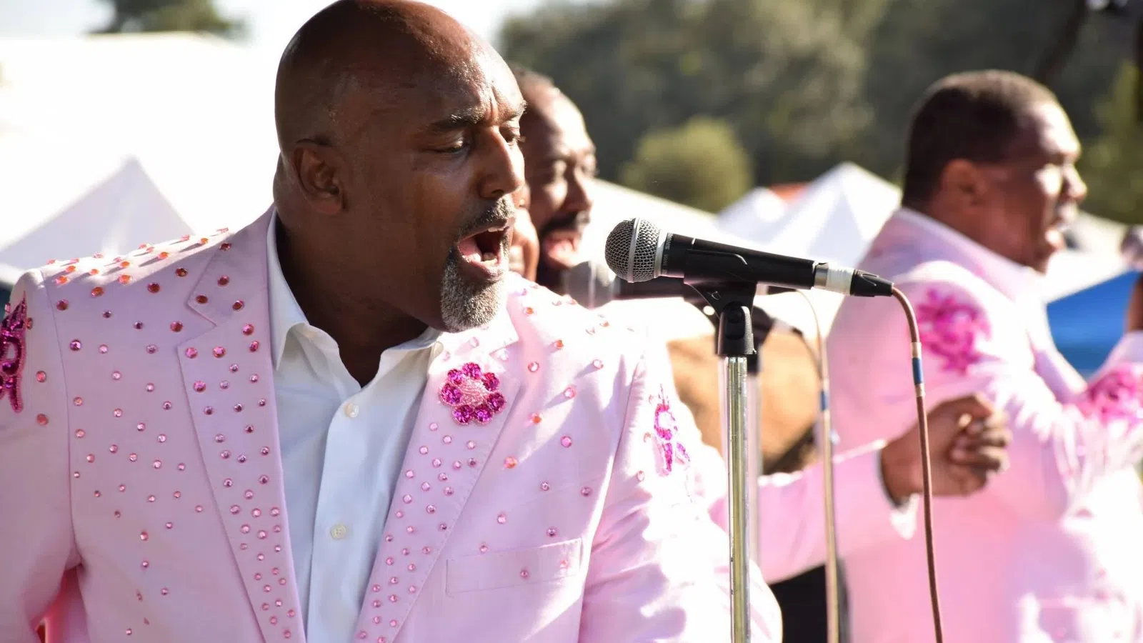 Male singers in matching pink embellished suits performing passionately at an outdoor event with microphones.
