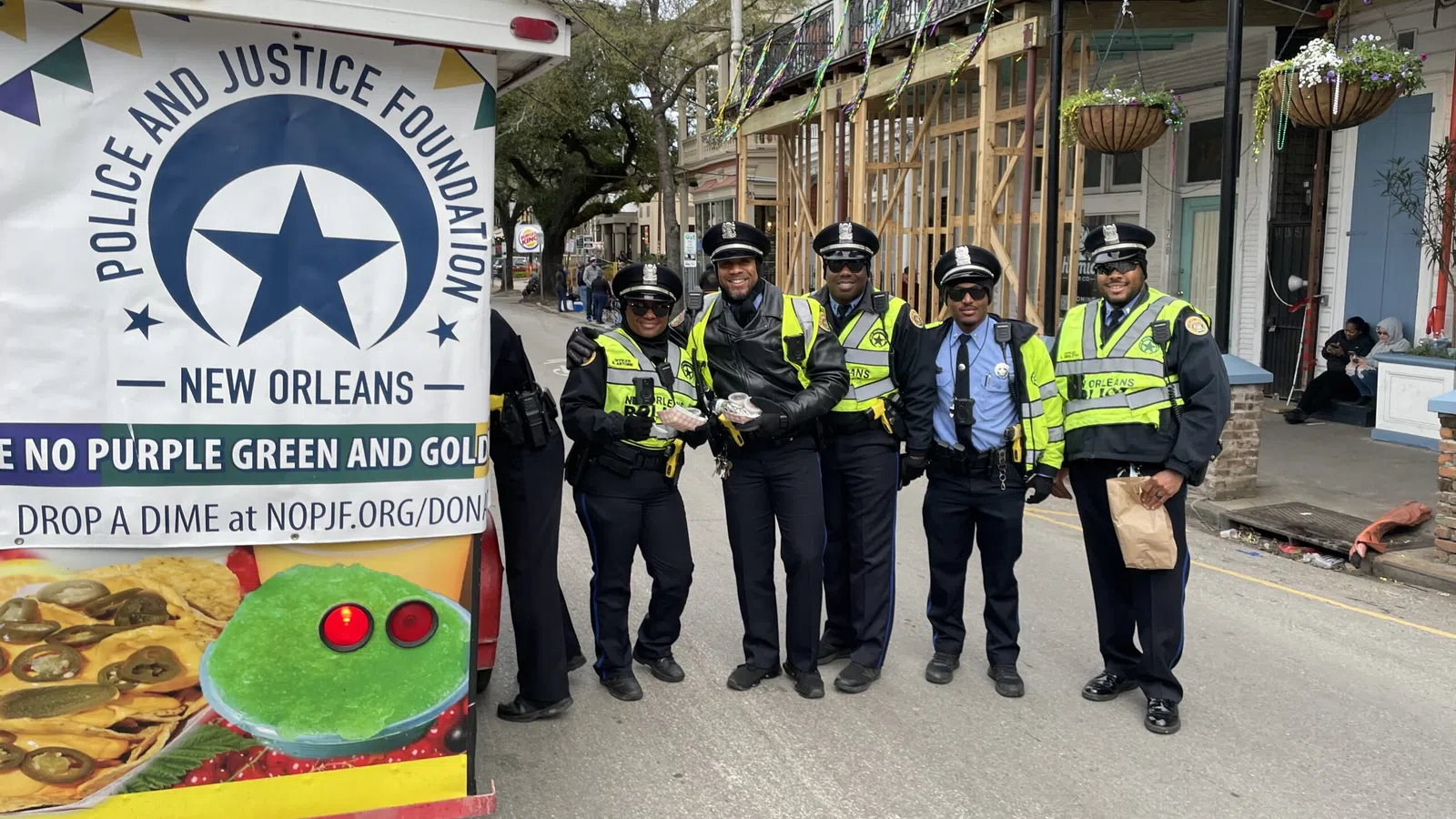 Five New Orleans police officers standing on a street beside a Police and Justice Foundation sign and Mardi Gras decorations.