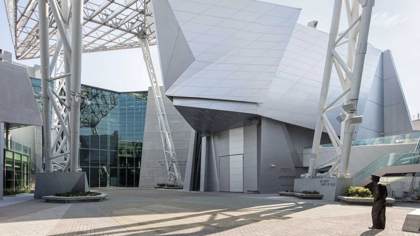 Modern architectural building with angular metal panels and large steel supports casting shadows on a paved courtyard.