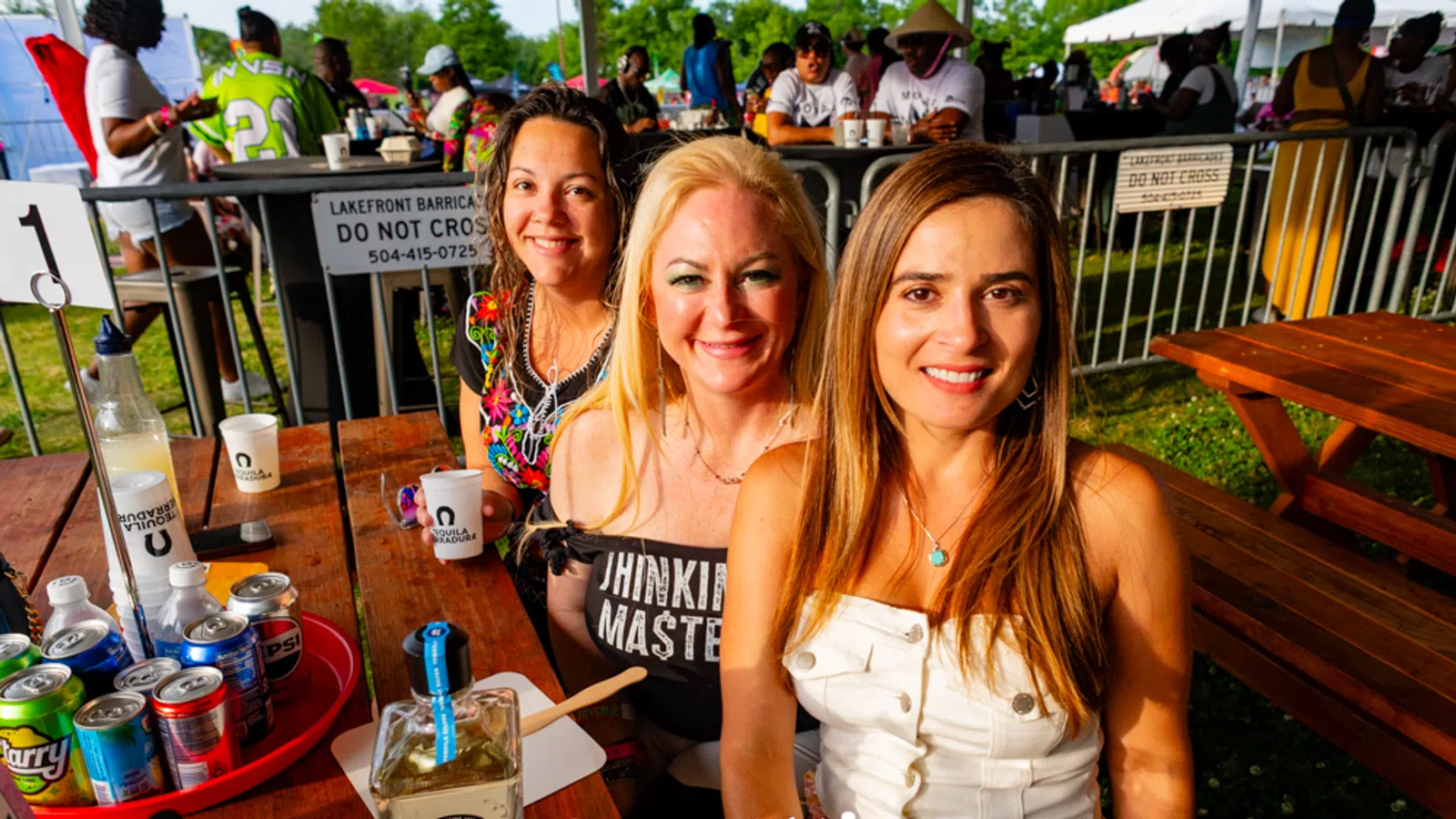 Three smiling women sitting at a wooden picnic table at an outdoor festival with drinks and beverages visible.