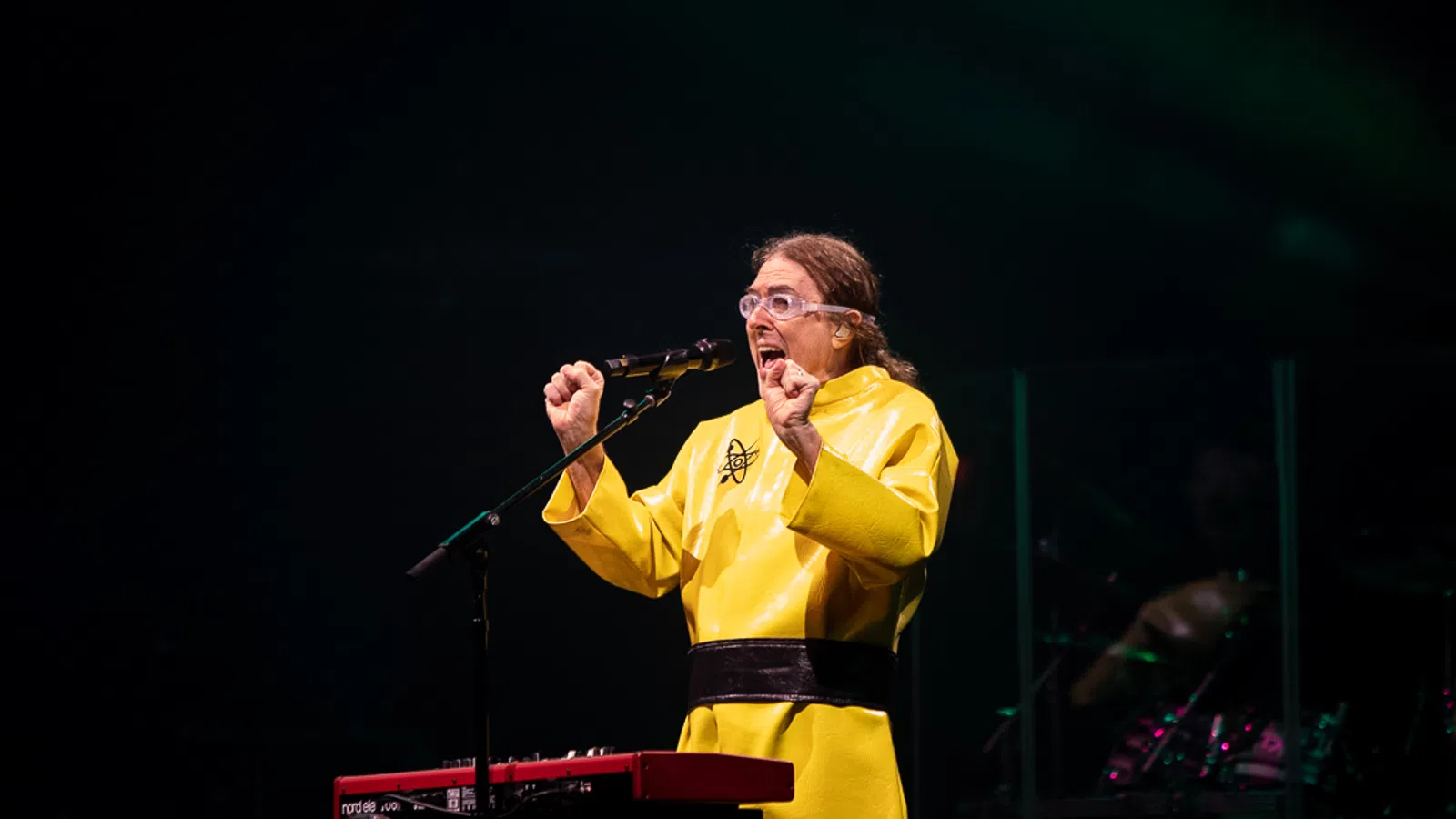 Performer in yellow costume sings passionately at microphone with keyboard and drum set in background on stage