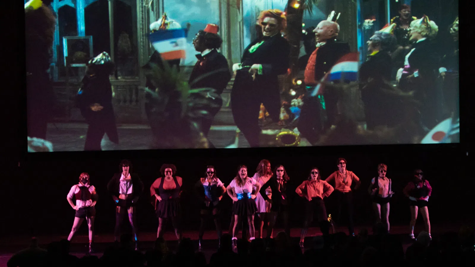 Performers in dark outfits stand on stage with a colorful projected backdrop during a theatrical show.