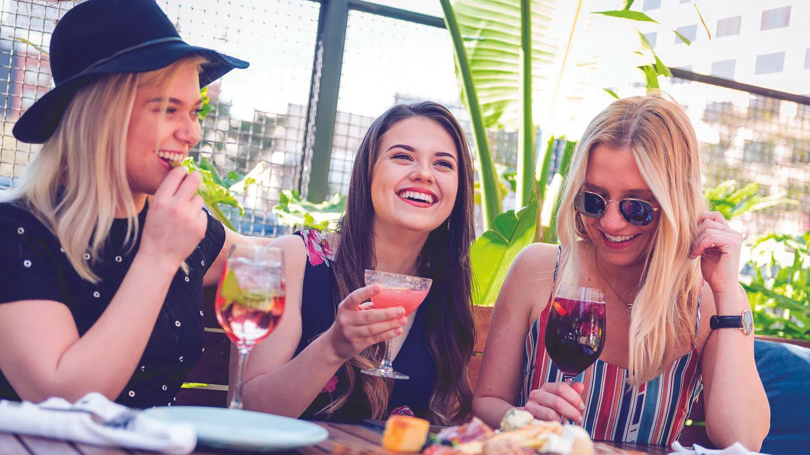 Three young women enjoy drinks and appetizers at an outdoor restaurant on a sunny day