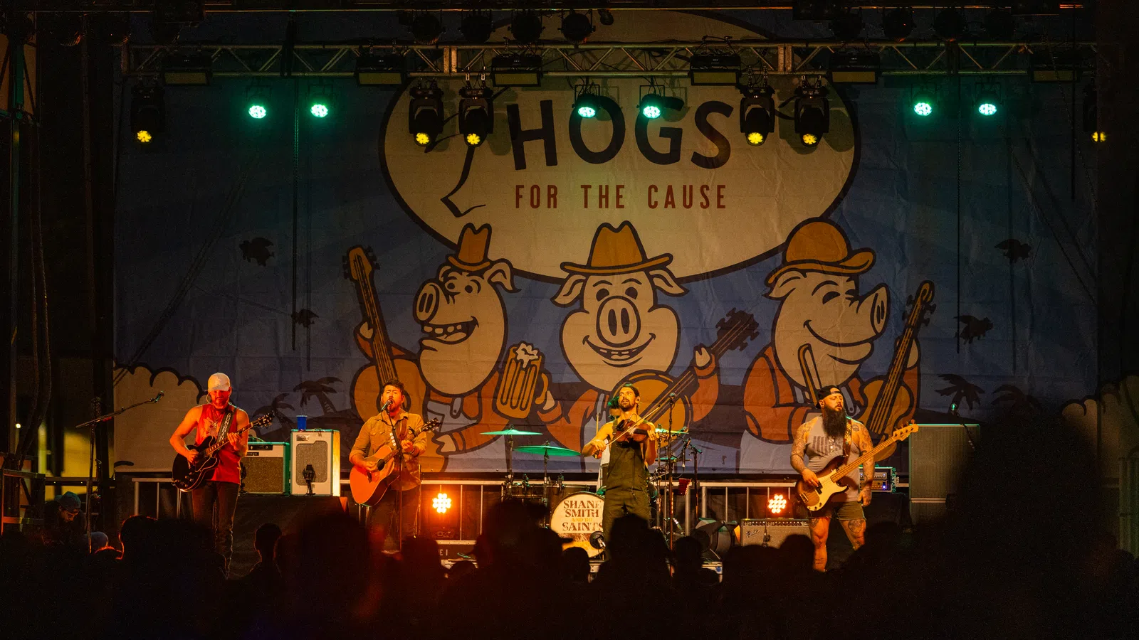 Band performing live on stage at night with Hogs for the Cause banner and cartoon pigs in background.