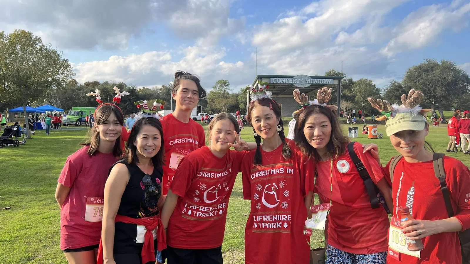 Group of seven people wearing festive red shirts and reindeer antlers posing outdoors at a park event on a sunny day